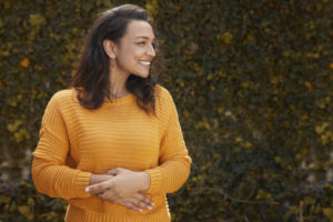 Portrait of beautiful happy woman standing infront of leafy wall with hands on stomach on a lovely autumn day