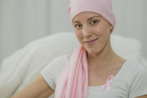 A woman with breast cancer is wearing a pink ribbon on her blouse and has a pink bandana around her head. She is hopeful and is smiling while looking at the camera.